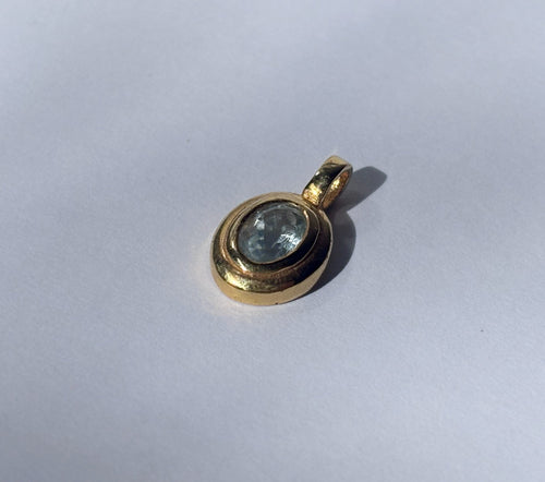 A close-up top-down view of a single, oval faceted pale blue aquamarine gemstone meticulously set within a simple, polished brass bezel on a white background. The pendant features a single brass loop for a chain