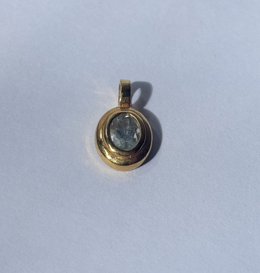 An angled close-up view of the pale blue aquamarine and polished brass pendant on a white background, showcasing the facet cut of the oval stone and the smooth bezel setting. The light creates dimensional qualities