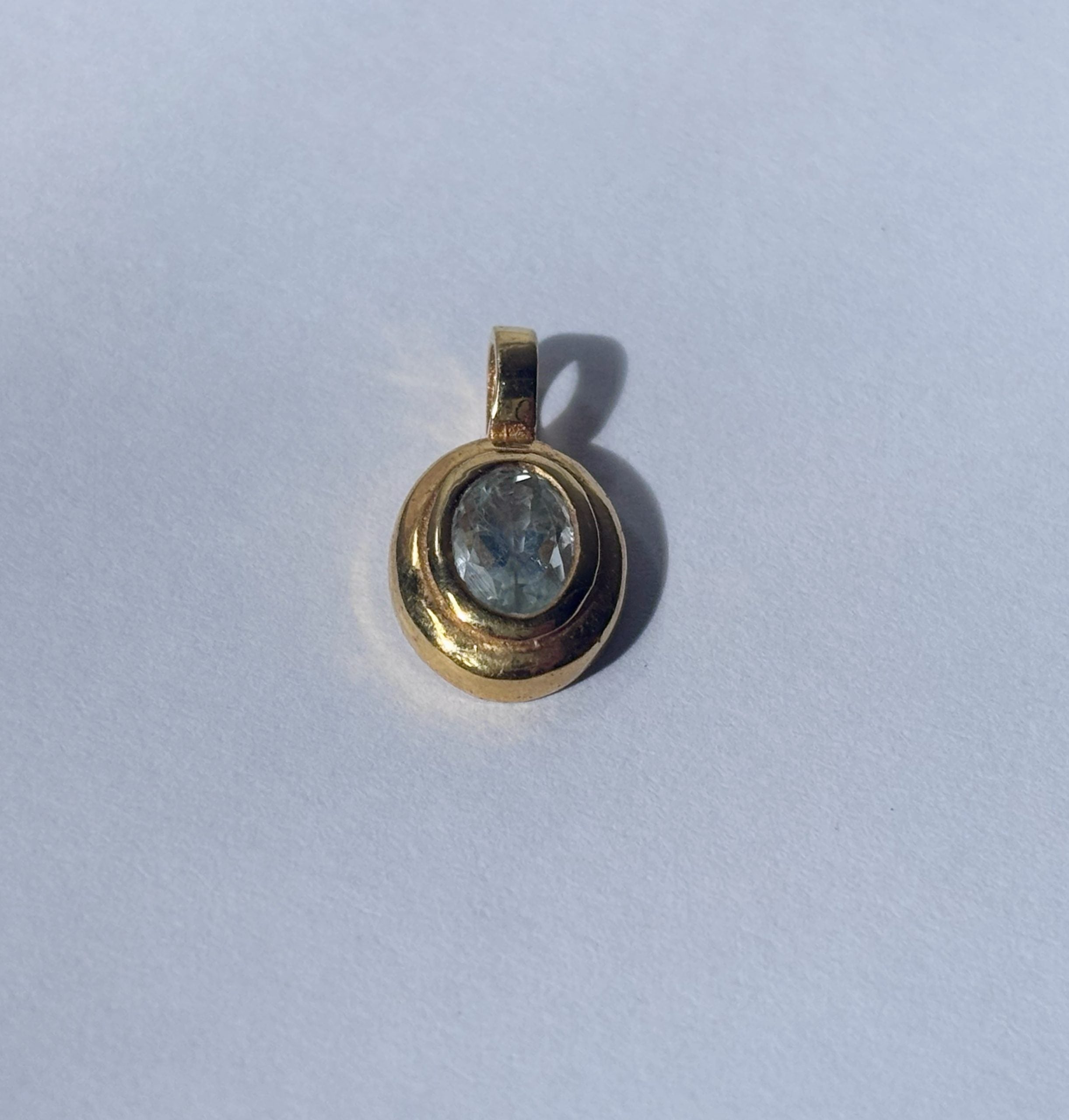 An angled close-up view of the pale blue aquamarine and polished brass pendant on a white background, showcasing the facet cut of the oval stone and the smooth bezel setting. The light creates dimensional qualities