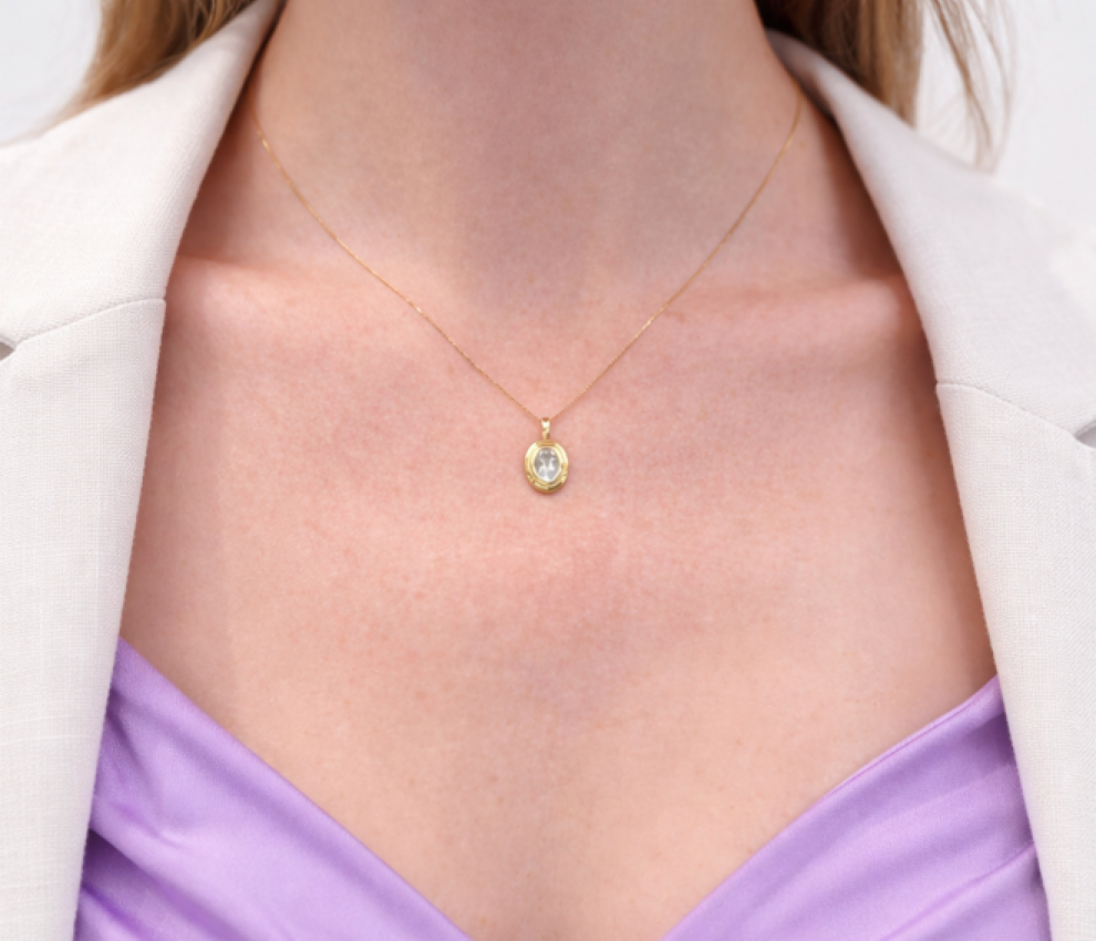 A minimalist everyday necklace featuring an oval faceted pale blue aquamarine gemstone pendant in a polished brass bezel setting, worn by a model over a purple top and white blazer. A delicate gold-tone chain is visible